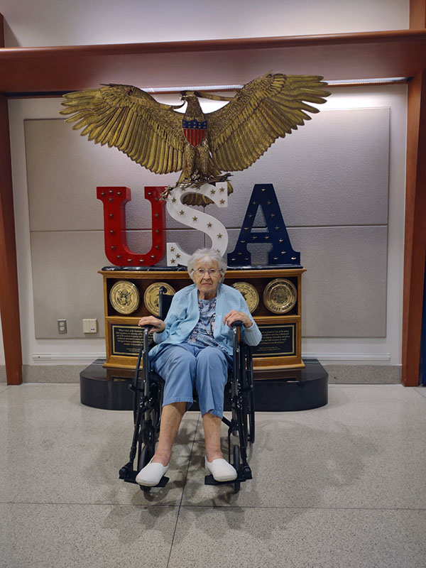Mrs. Josephine Ann Talipin Gazelle in her wheelchair posing in front of a statue of a large American bald eagle which is sitting atop the red, white, and blue colored letters 'USA'